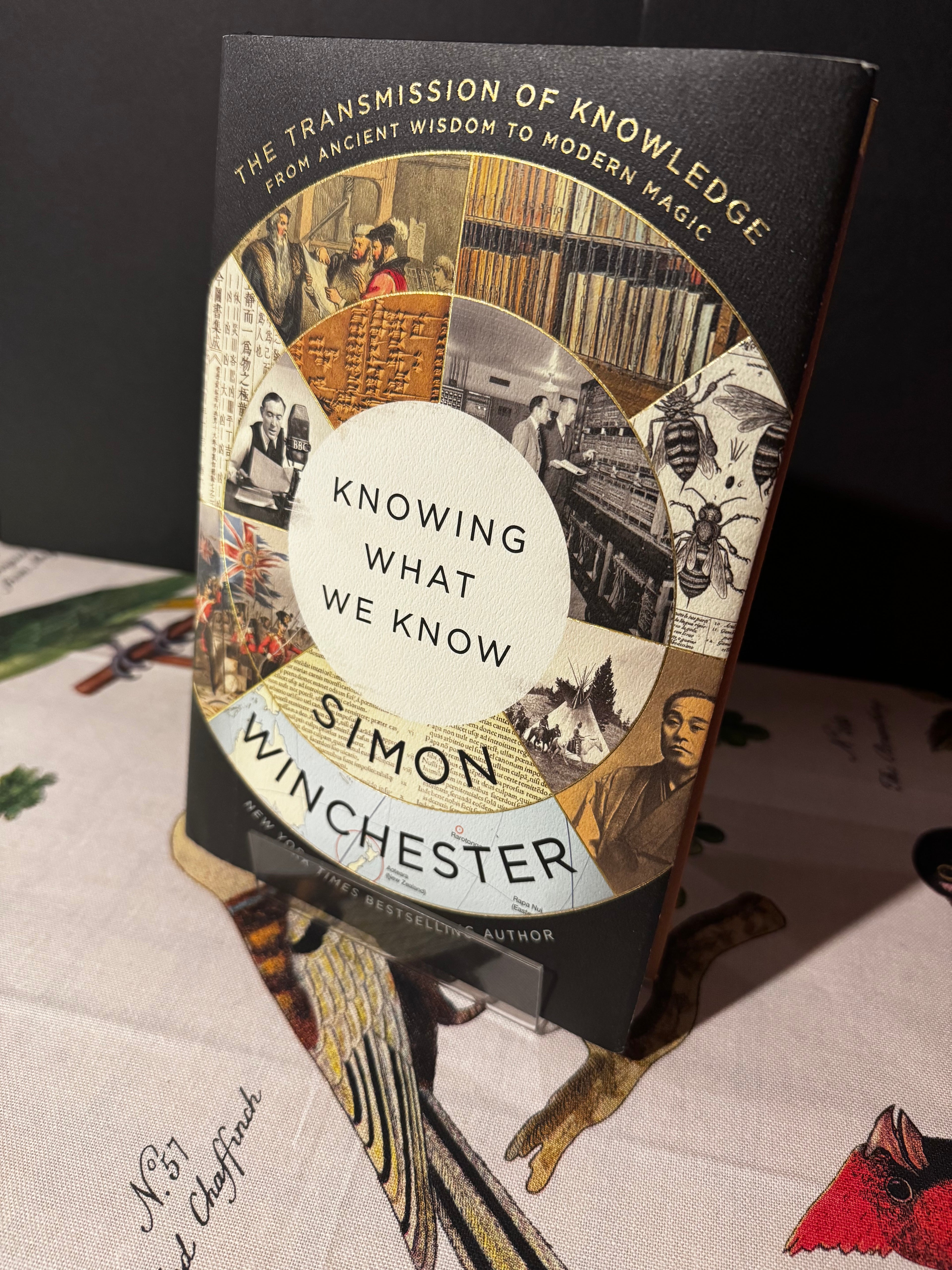 Book titled 'Knowing What We Know' by Simon Winchester on a textured surface with artistic elements.