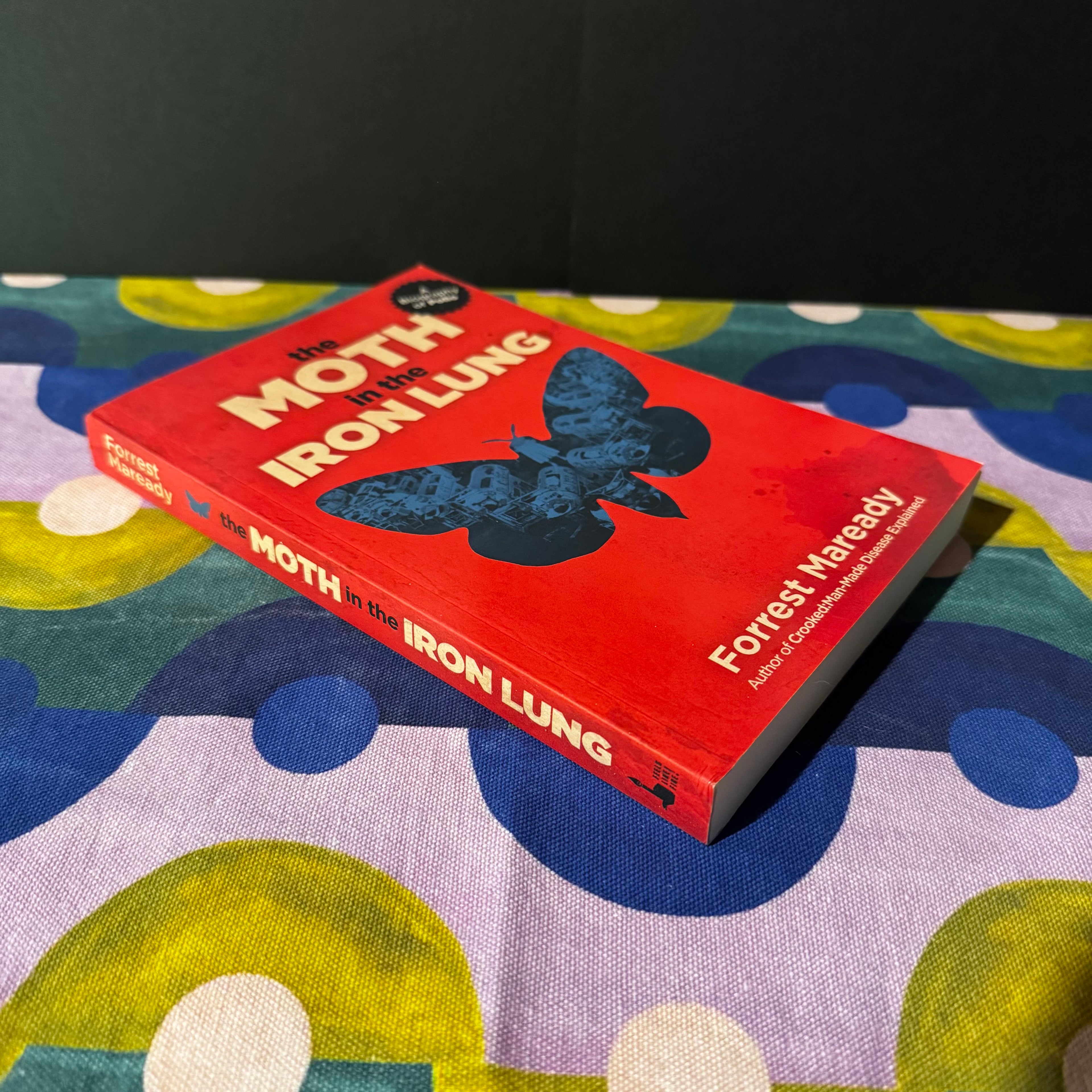 Book titled 'The Moth in the Iron Lung' by Forrest Maready on a colorful fabric background