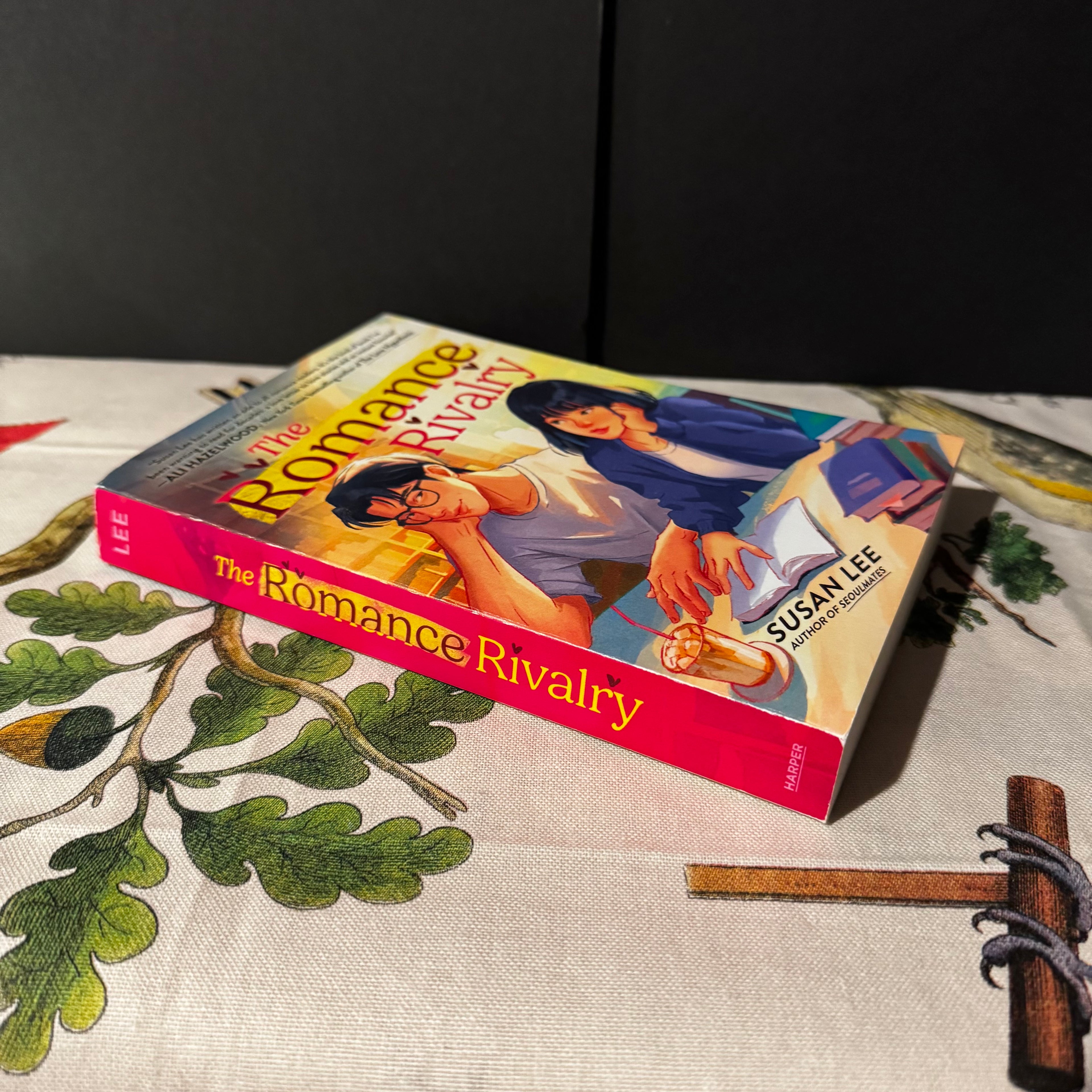 Book titled 'The Romance Rivalry' by Susan Lee on a decorative surface with leaves and twigs.