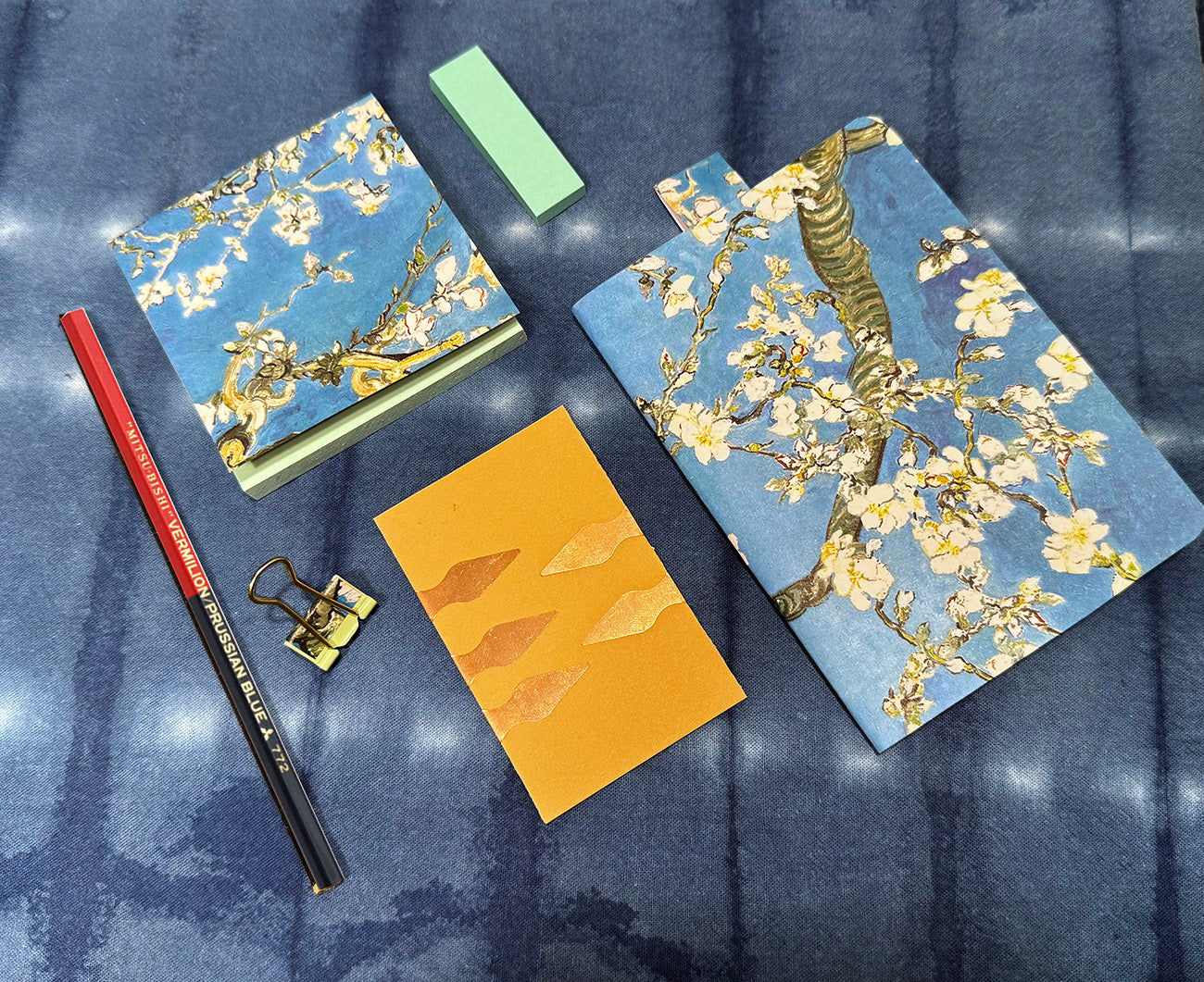 Two notebooks with floral design on a textured blue surface, with a pencil and clip on top.