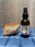 Brown spray bottle labeled 'Area Spray' next to a crystal on a blue surface with a gray textured background