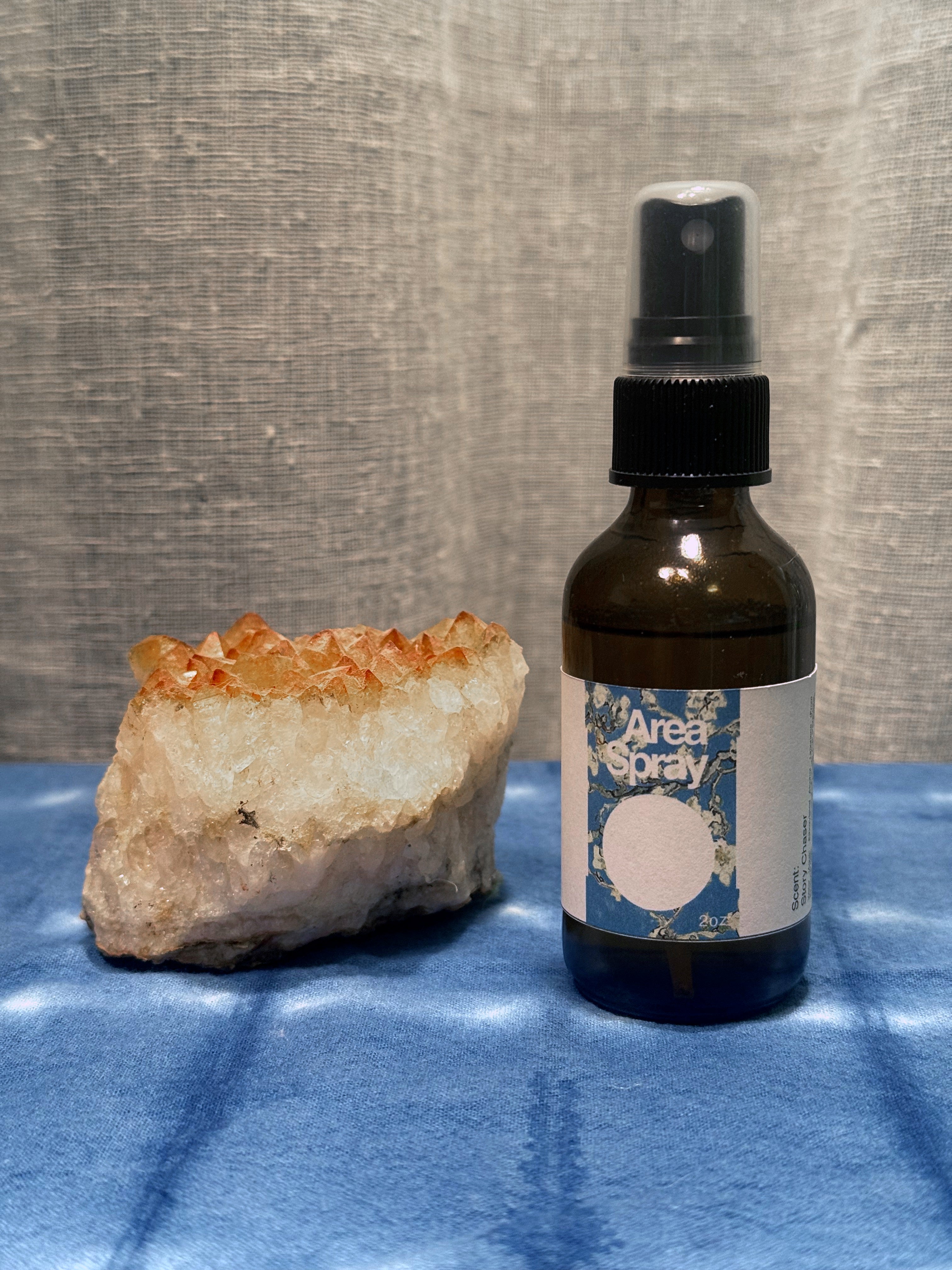 Brown spray bottle labeled 'Area Spray' next to a crystal on a blue surface with a gray textured background